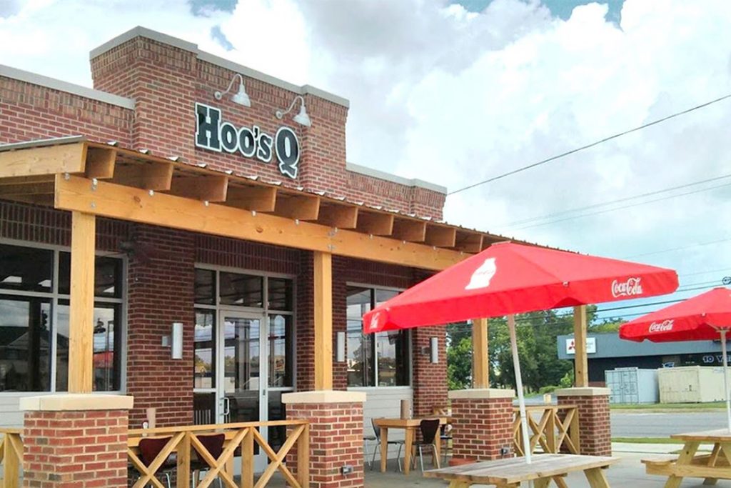 A brick building with a wooden overhang and Coca-Cola umbrellas, featuring outdoor seating and a sign reading 'HoosQ' in Tuscaloosa, Alabama.