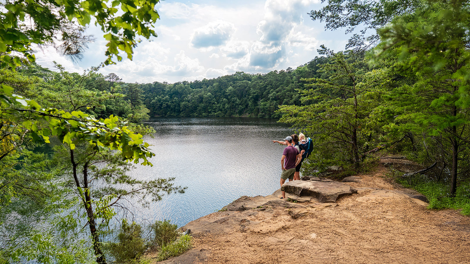 A group of people standing on an overlook to a lake with one person pointing to something in the distance.