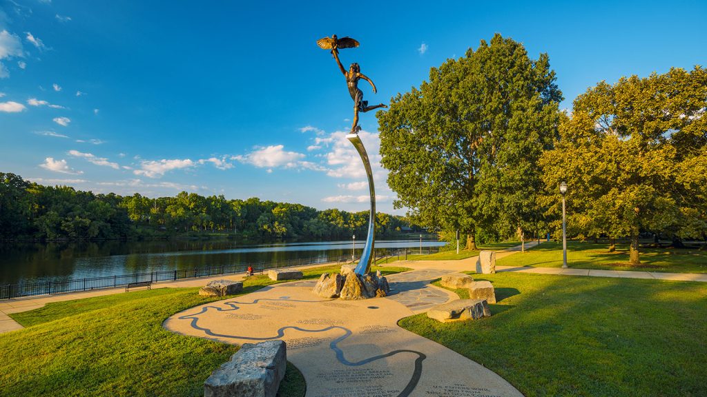 A statue of a person holding a bird by the feet with a view of the sky and lake in the background.