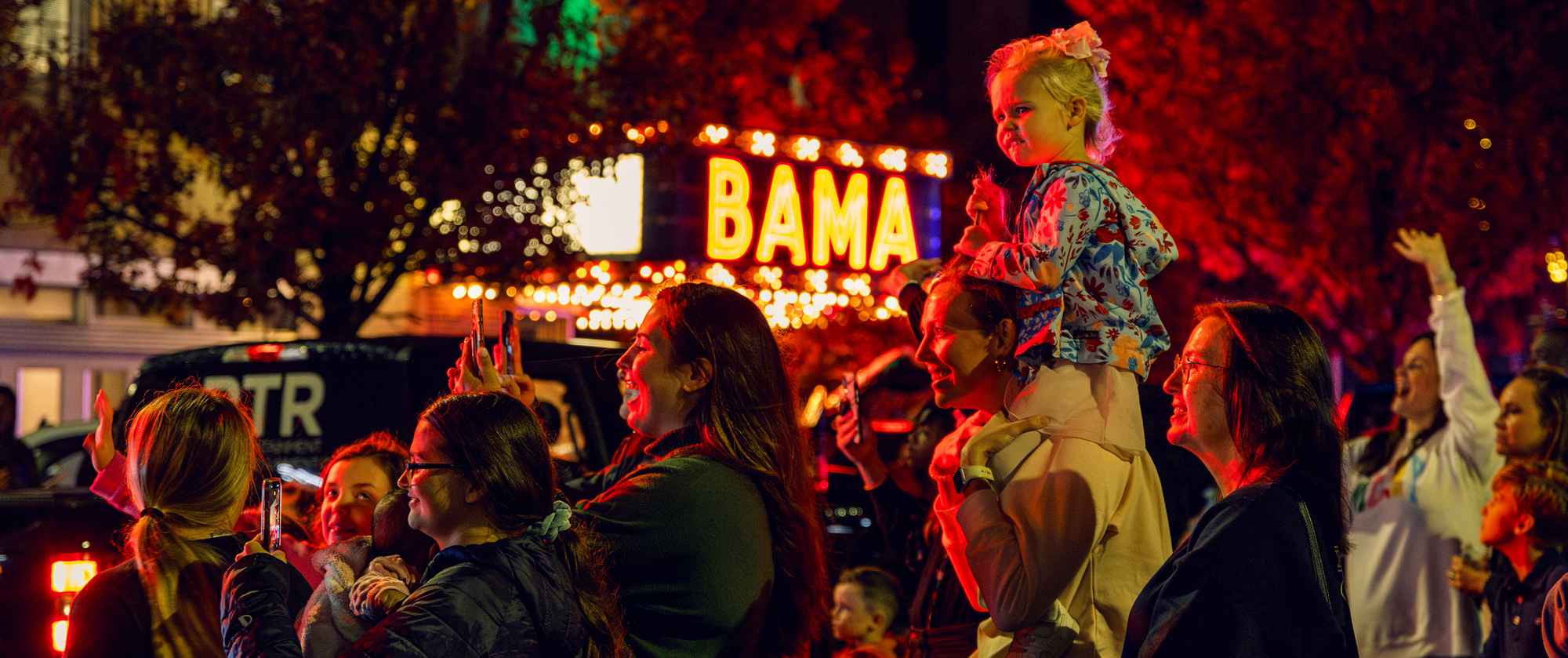 A crowd of people with a child sitting on a person's shoulders with a theater marquee in the background.