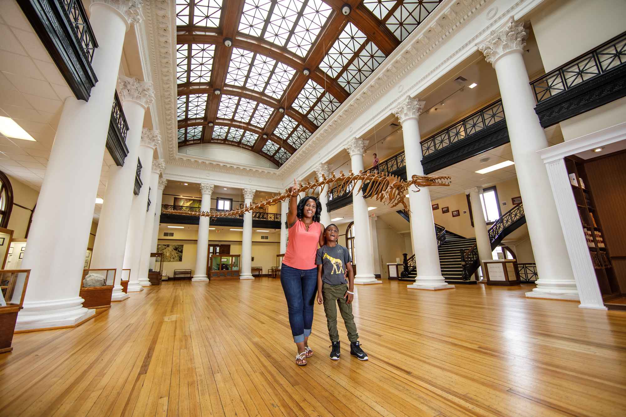 A mother and child explore the AL Museum of Natural History. A dinosaur skeleton is seen in the background.