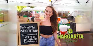 A woman stands at a Margarita Festival booth, holding a glass of margarita and smiling next to a sign advertising a margarita competition