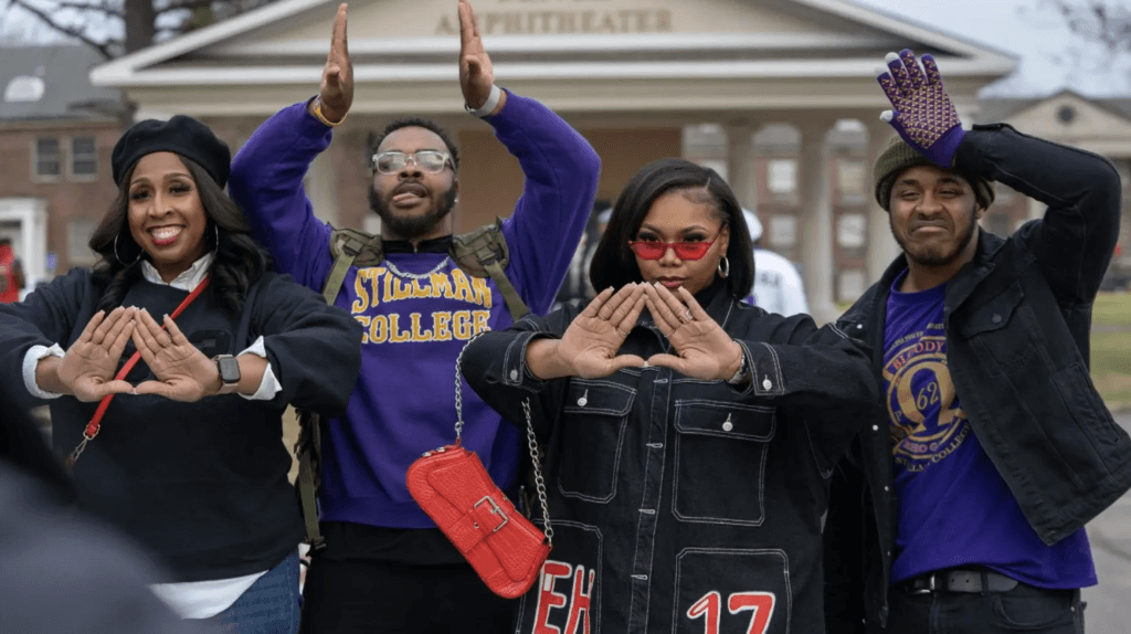 Four individuals, dressed in casual attire with Greek letters, stand in front of a building, making hand gestures associated with fraternities or sororities