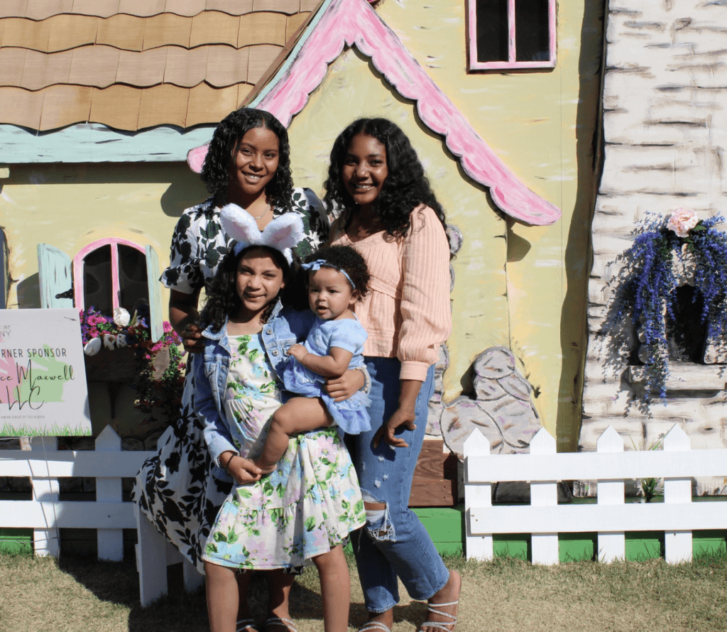 Four women and a baby pose in front of a whimsical, pastel-colored house with a thatched roof and floral decorations