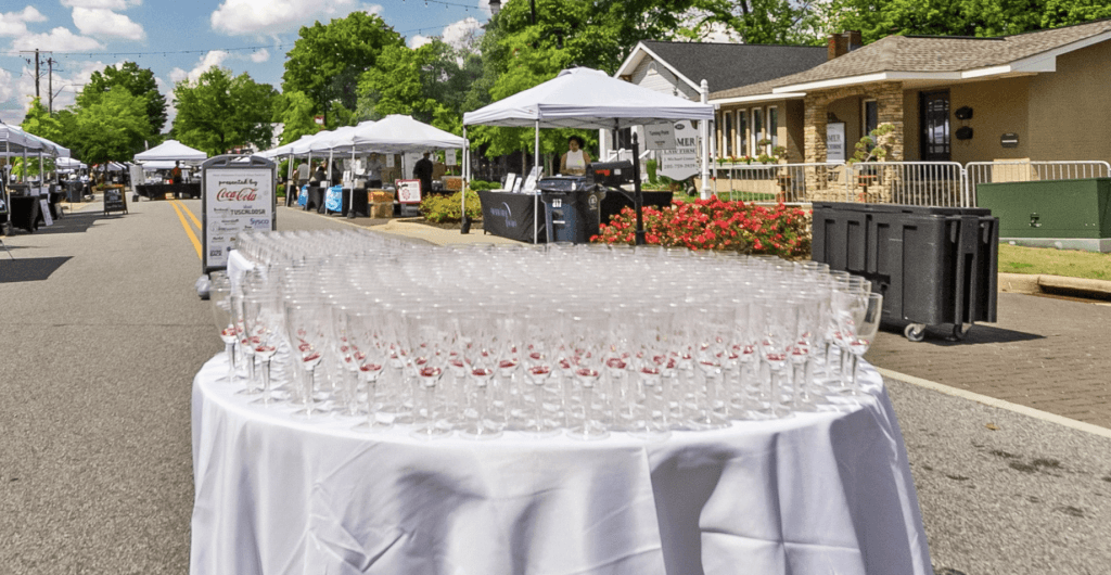A long table covered with a white tablecloth is set up on a street, laden with neatly arranged rows of empty wine glasses