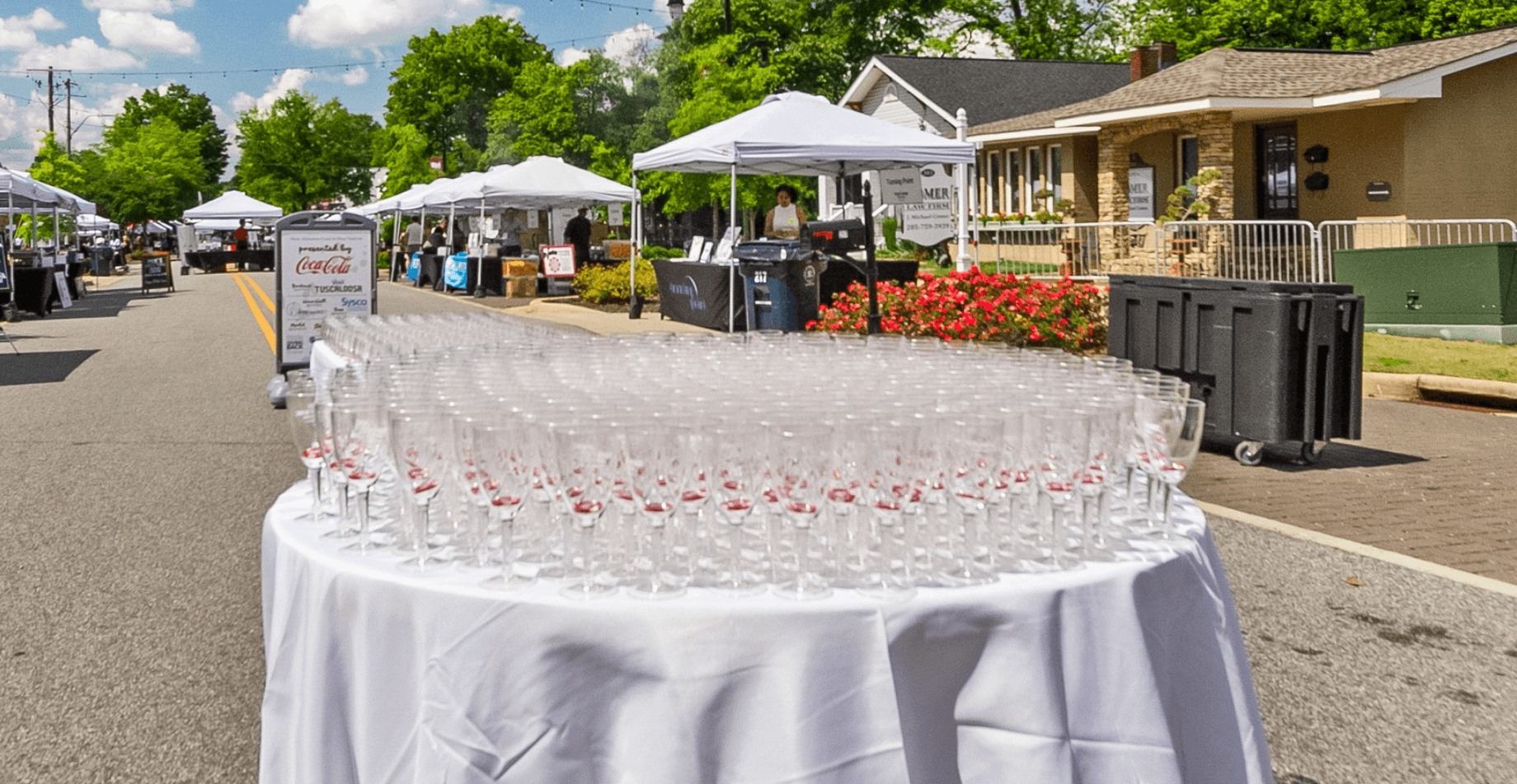 A long table covered with a white tablecloth is set up on a street, laden with neatly arranged rows of empty wine glasses