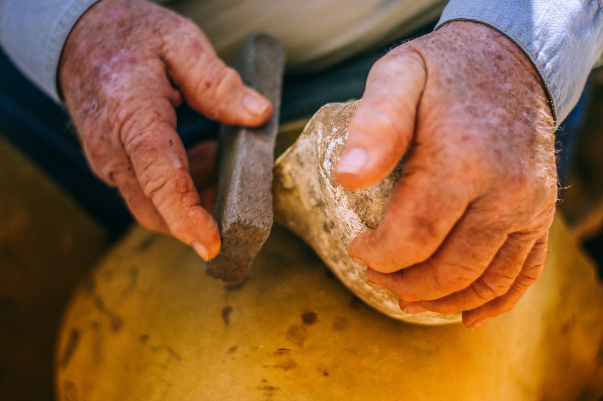 An elderly person's weathered hands carefully shape a piece of clay using a stone tool, demonstrating traditional pottery techniques