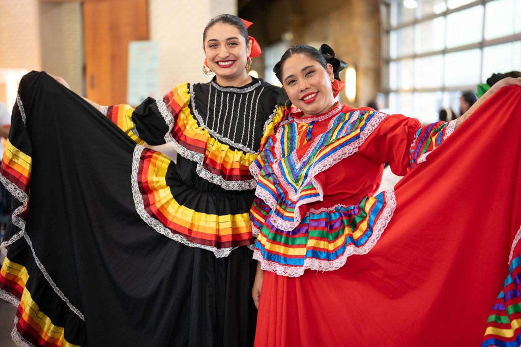Two women in vibrant traditional Mexican dresses pose with their skirts spread wide, showcasing colorful ruffles and intricate lace details