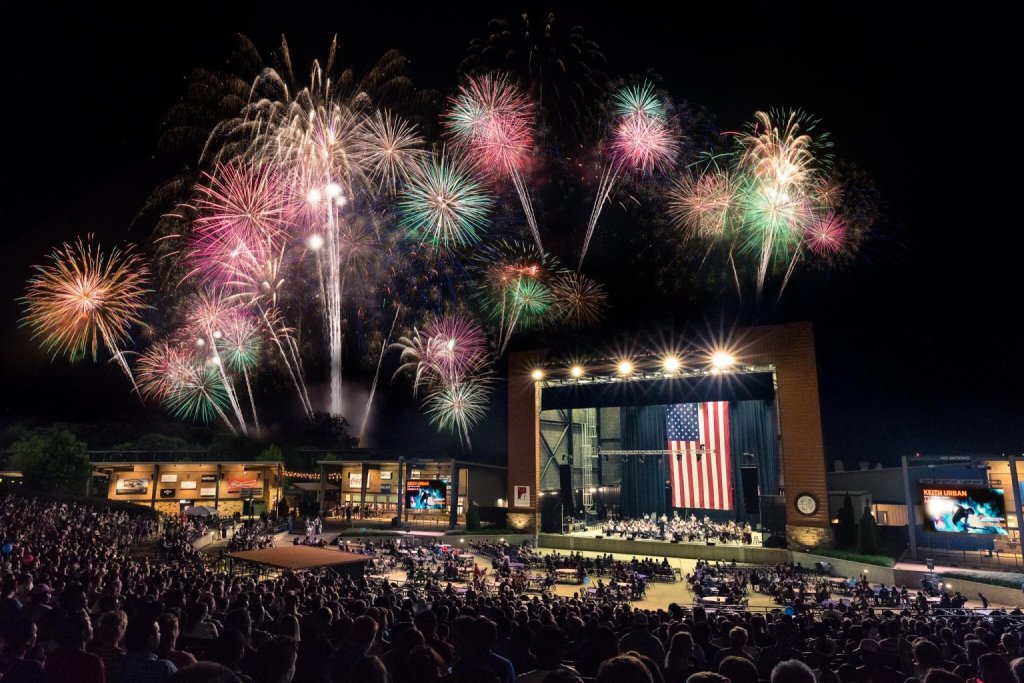A vibrant fireworks display illuminates the night sky above a packed outdoor concert venue, with a large American flag hanging prominently on stage