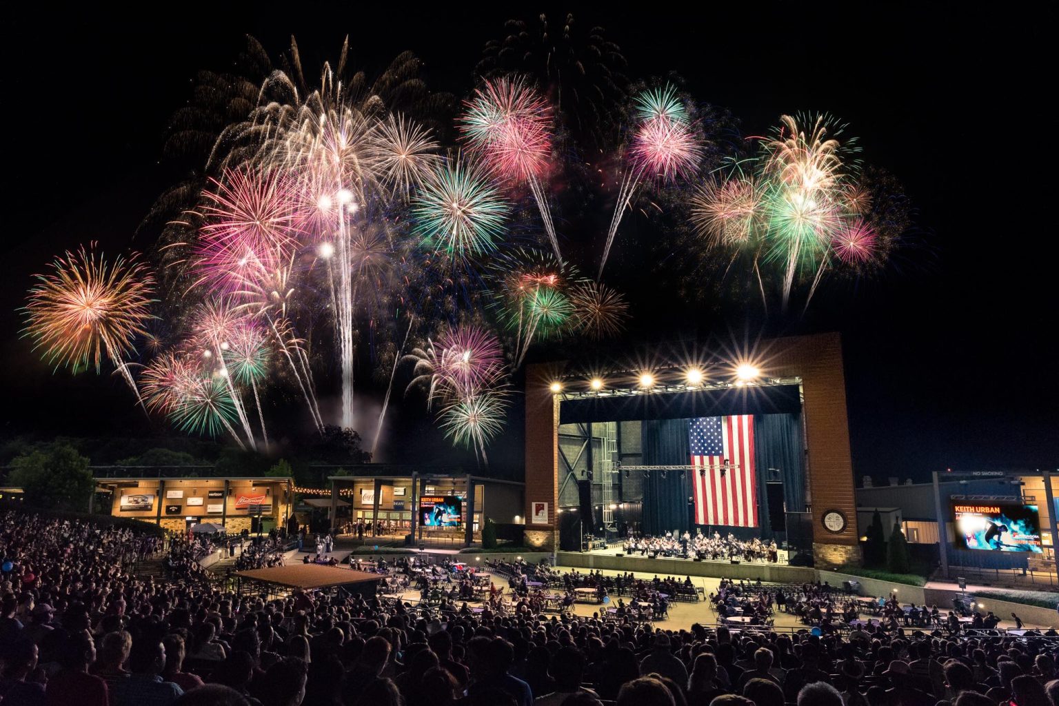 A vibrant fireworks display illuminates the night sky above a packed outdoor concert venue, with a large American flag hanging prominently on stage