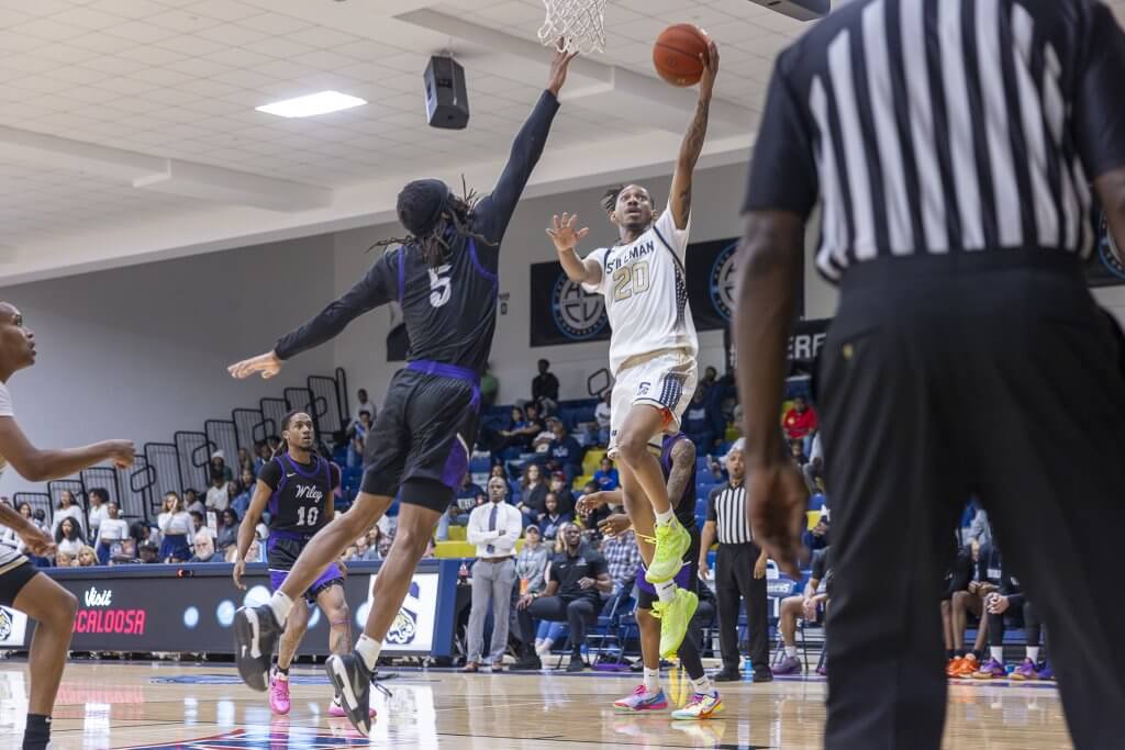 A basketball player in a white jersey with the number 20 leaps high to dunk the ball, while a defender in a black jersey with the number 5 tries to block the shot
