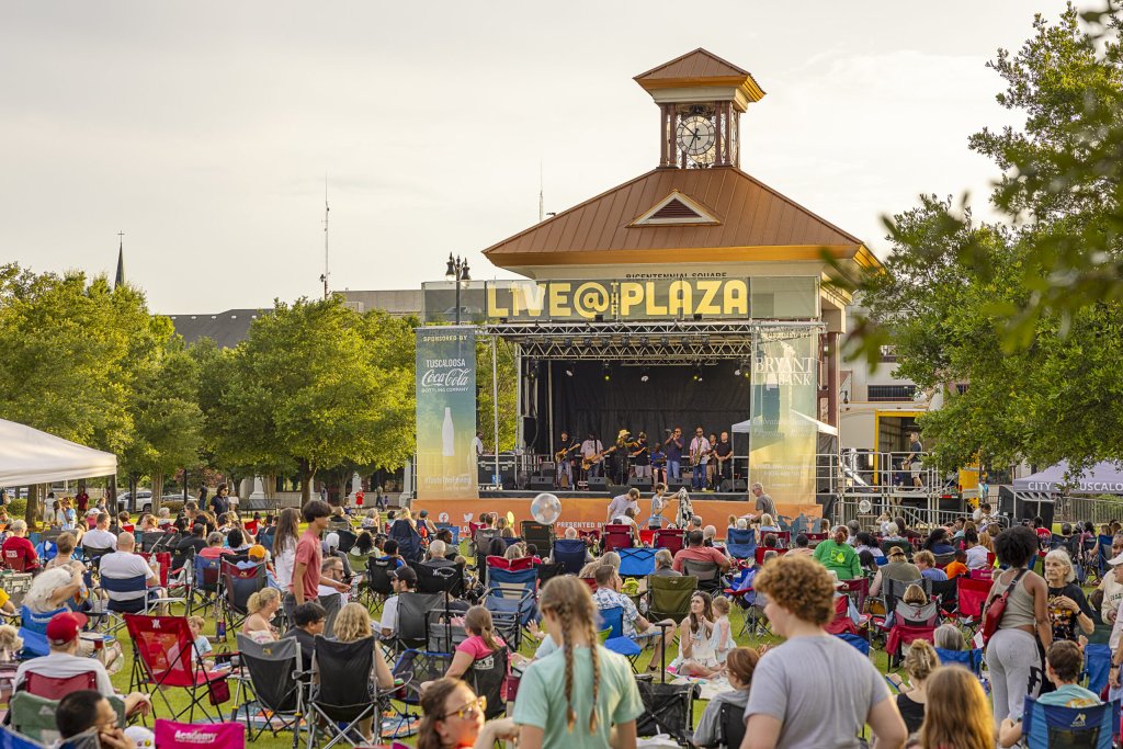 A lively outdoor concert at Plaza Live features a diverse crowd enjoying music under a clear sky, with a distinctive clock tower and vibrant stage setup in the background