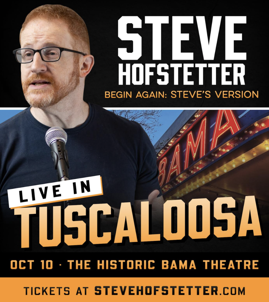 Steve Hofstetter, a red-haired man with glasses, speaks into a microphone on stage at the Historic Bama Theatre in Tuscaloosa, Alabama