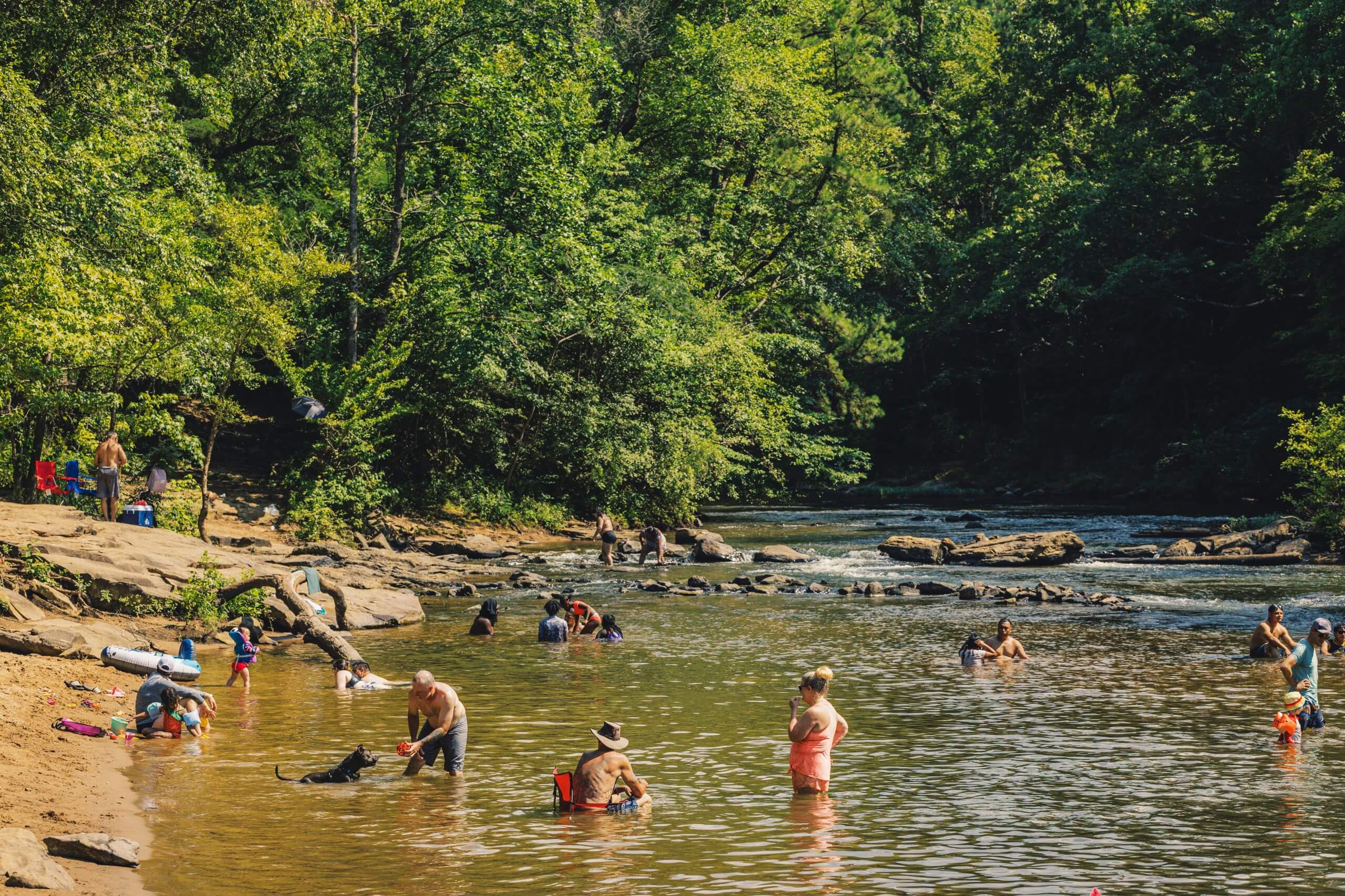 A lively scene unfolds at a riverbank where numerous people are enjoying a sunny day, swimming, playing, and relaxing in the water and on the sandy shore, surrounded by lush greenery