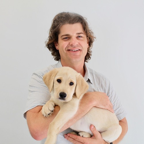 A man with curly hair is smiling as he holds a light-colored puppy close to his chest