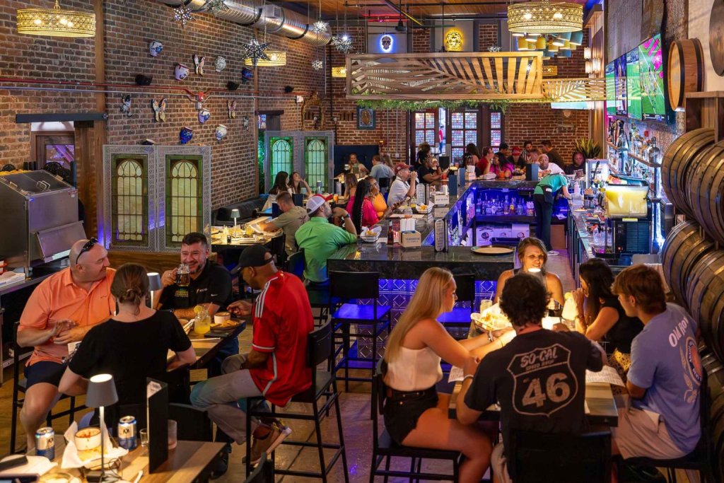A lively bar scene with patrons enjoying drinks and meals at tables and the bar, surrounded by eclectic decor and exposed brick walls