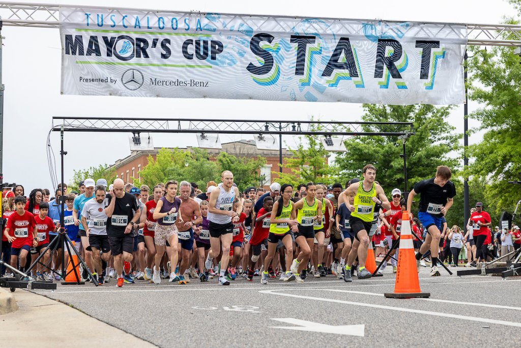 A bustling marathon start scene with runners gathered under a large banner at the beginning of the Tulsa Mayor's Cup