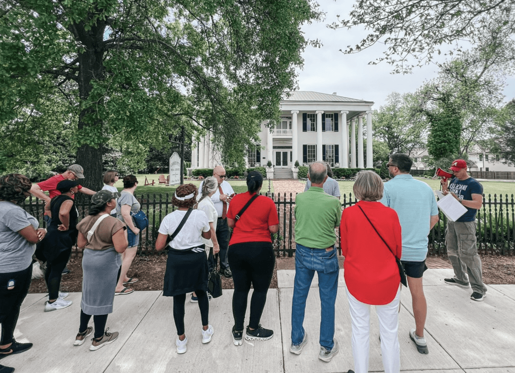 Tourists gather around a grand white mansion, listening to a guide's speech in Tuscaloosa, Alabama.