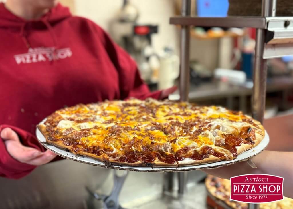 A person in a red hoodie holds a freshly baked pizza with melted cheese and toppings in Tuscaloosa, Alabama.