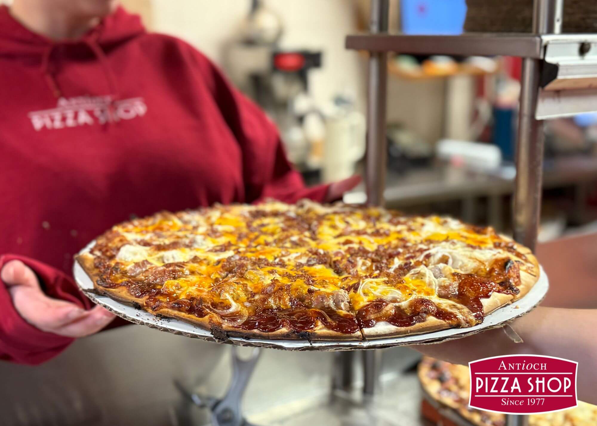 A person in a red hoodie holds a freshly baked pizza with melted cheese and toppings in Tuscaloosa, Alabama.