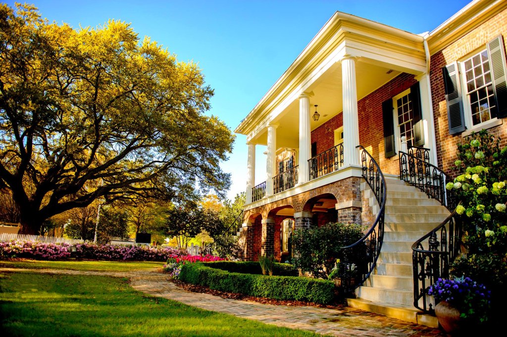 A grand, historic house with a white porch and black railings, surrounded by lush gardens and a large tree in Tuscaloosa, Alabama.