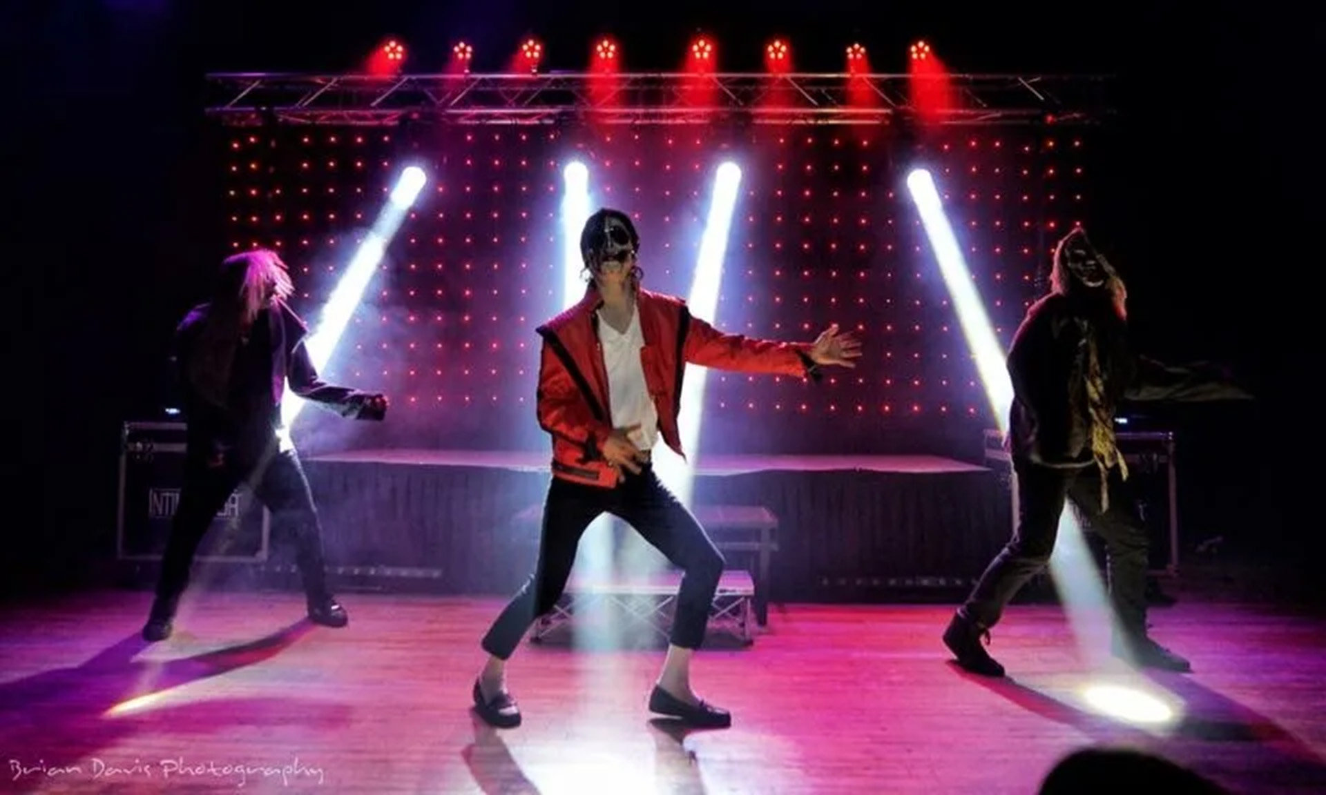Three performers on stage, one in red jacket, two in black, with red lighting and laser beams