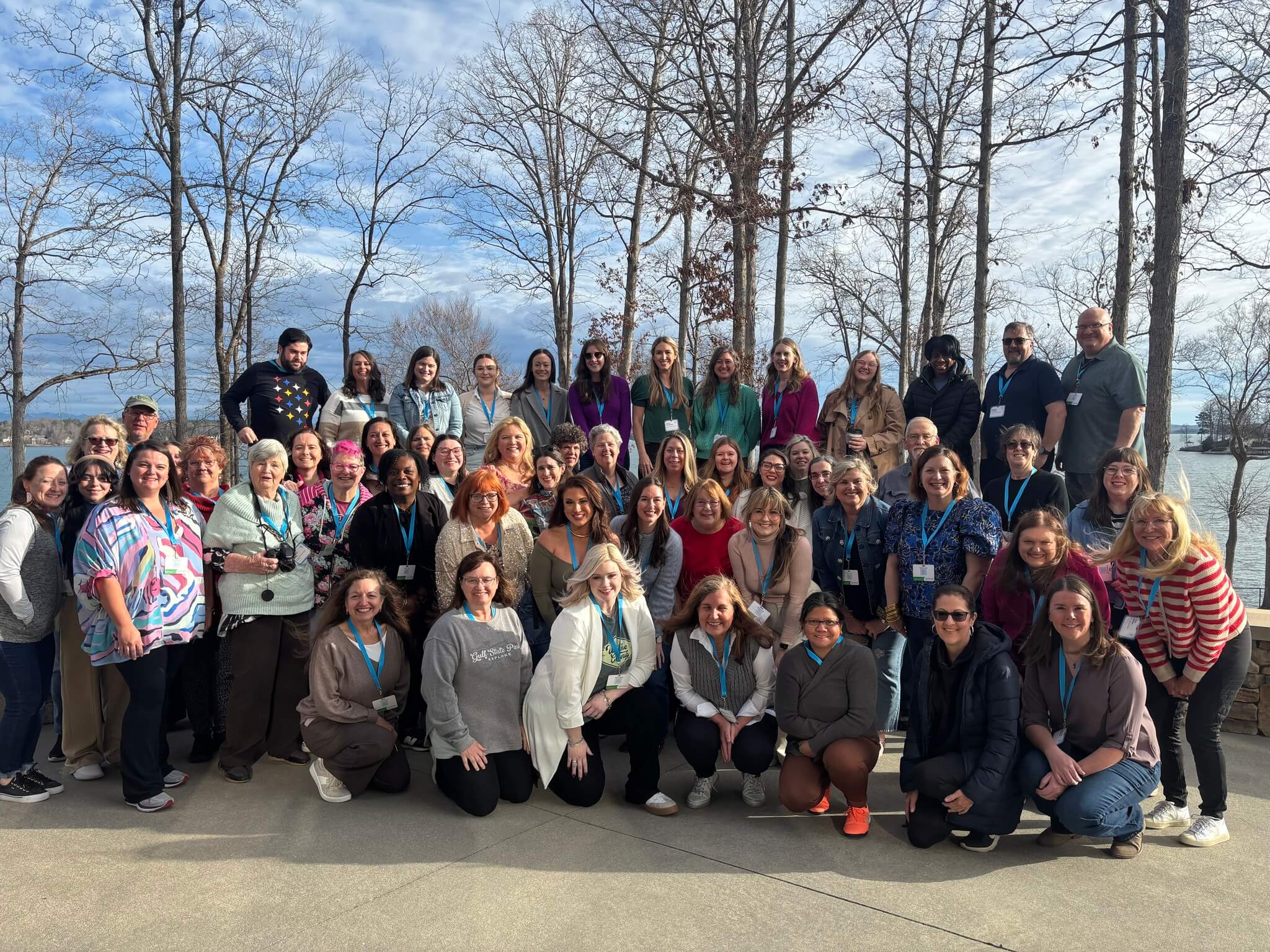 A diverse group of people, many wearing lanyards, gather for a group photo outdoors with trees and a lake in the background in Tuscaloosa, Alabama.