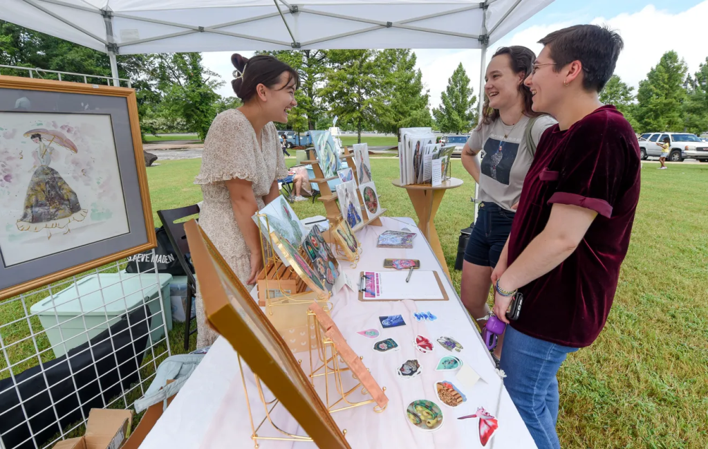Three women at an outdoor art fair, laughing and examining paintings in Tuscaloosa, Alabama.