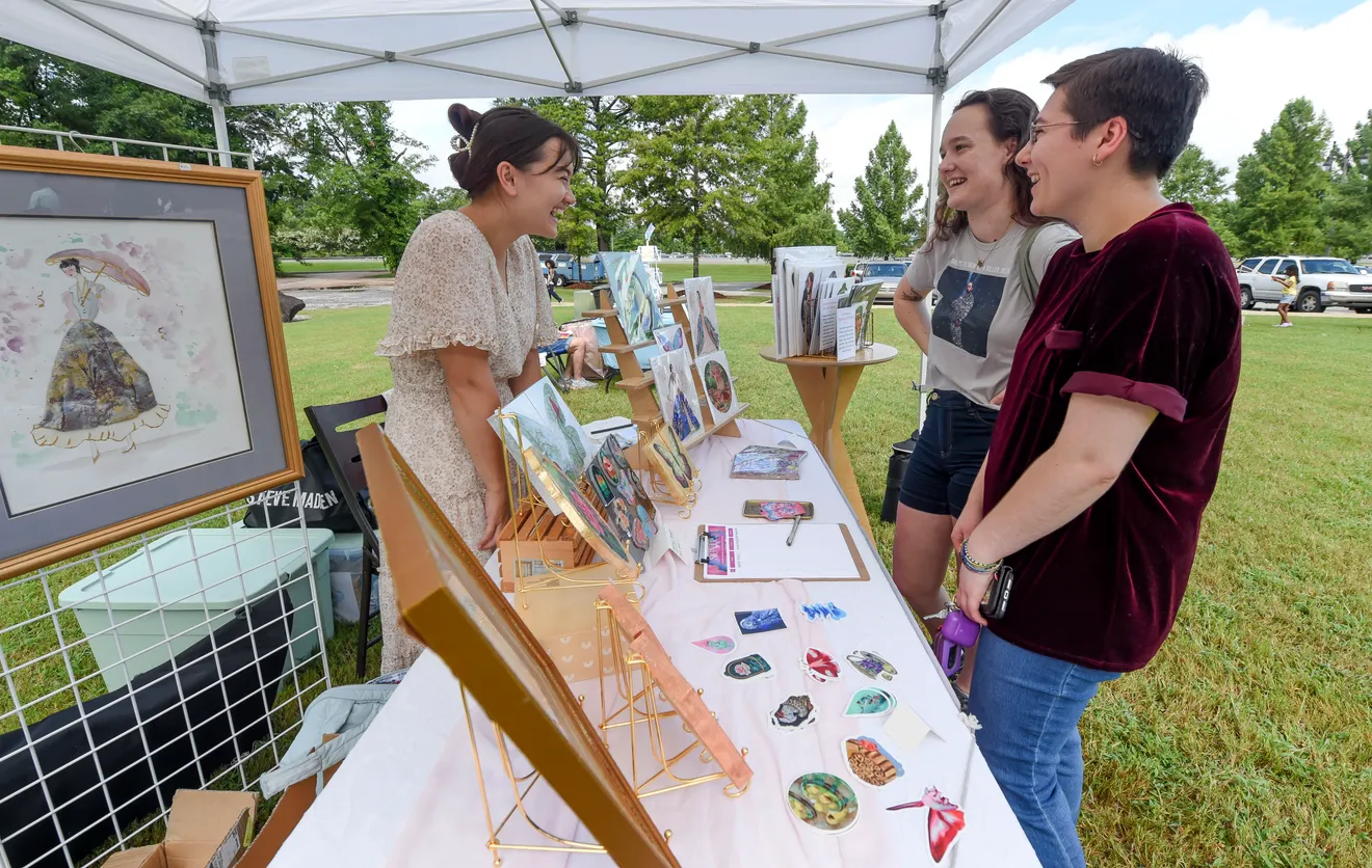 Three women at an outdoor art fair, laughing and examining paintings in Tuscaloosa, Alabama.