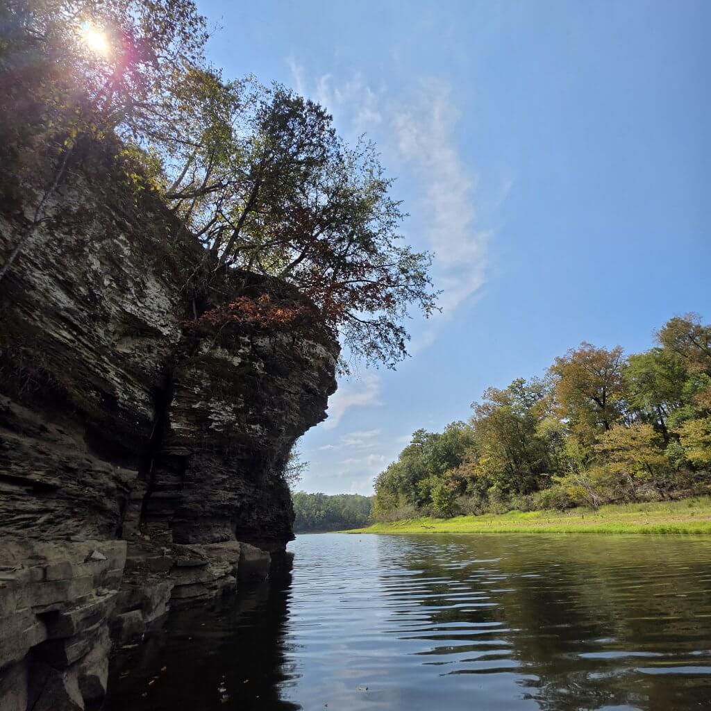 A serene river winds through a lush forest, with a towering rock cliff adorned with autumn foliage on one side and a clear blue sky above in Tuscaloosa, Alabama at Lake Nicol.