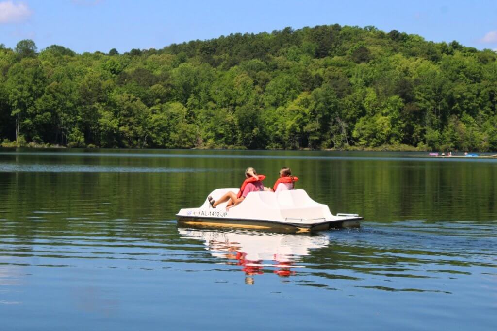 Two people wearing life jackets paddle a small boat on a serene lake surrounded by lush green trees in Tuscaloosa, Alabama at Lake Lurleen State Park.