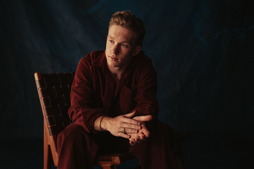 A young man in a red outfit sits thoughtfully on a wooden chair, with a dark background in Tuscaloosa, Alabama.
