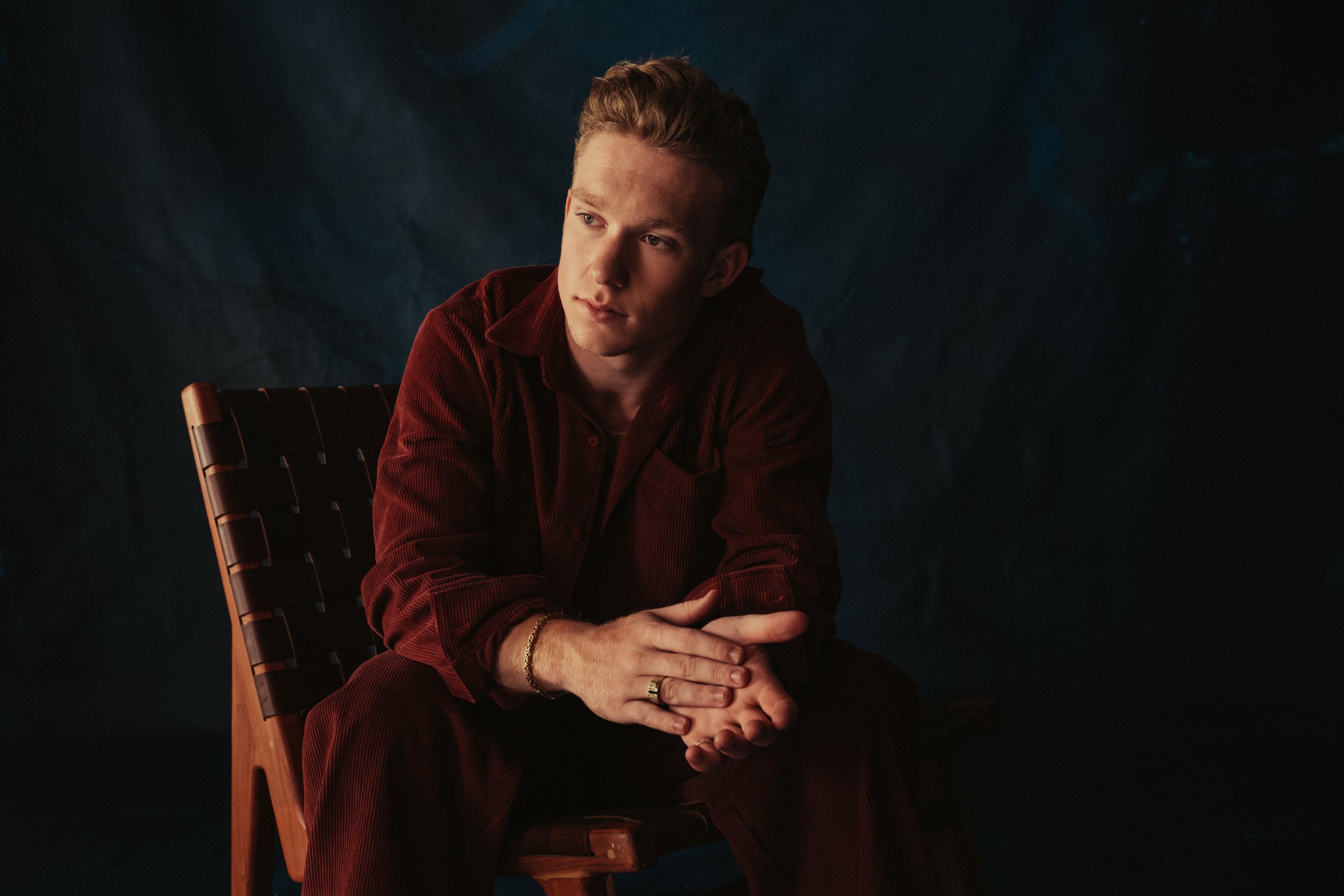 A young man in a red outfit sits thoughtfully on a wooden chair, with a dark background in Tuscaloosa, Alabama.