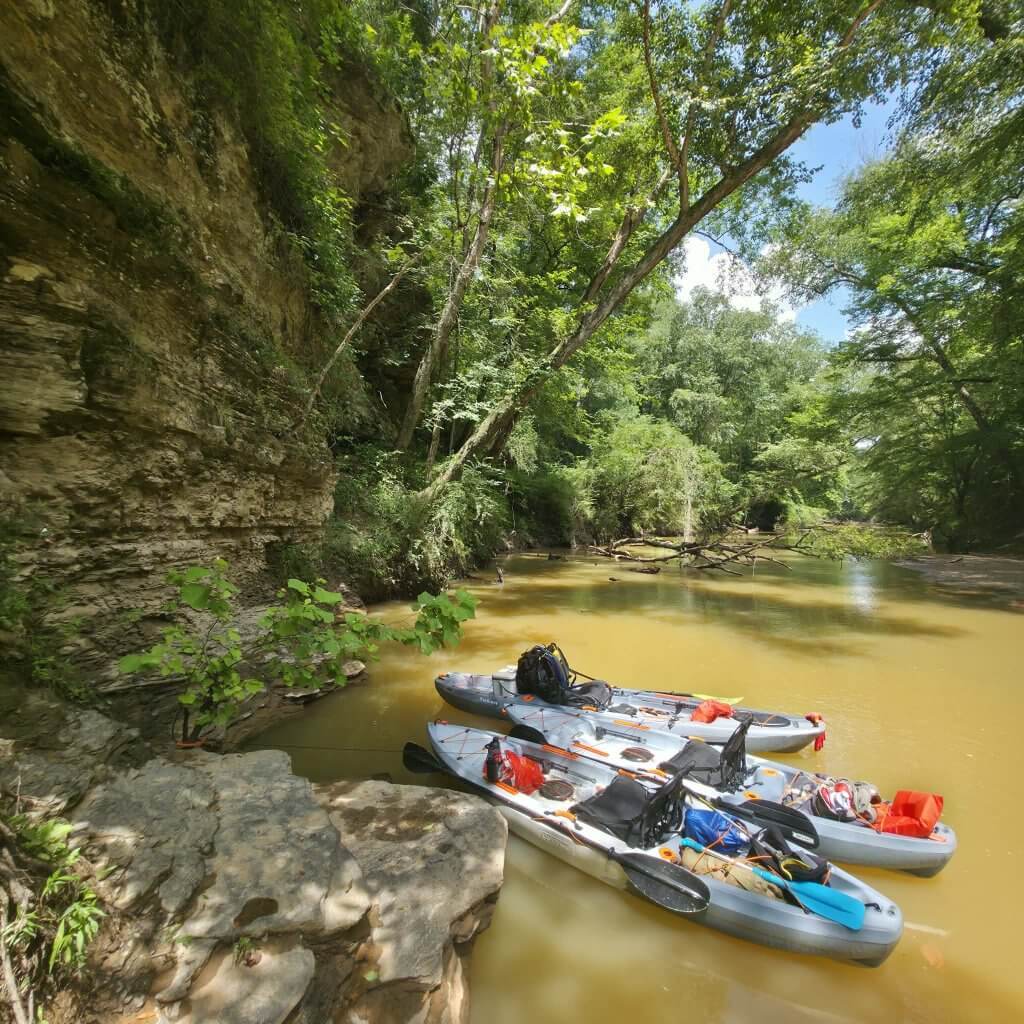 Two kayaks are moored to a rock near a muddy riverbank, surrounded by lush green trees and a towering cliff in Tuscaloosa, Alabama along Hurricane Creek.