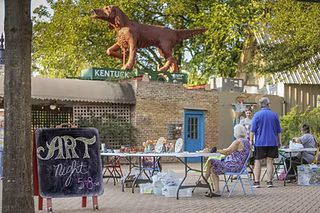 A vibrant art night with a large dog statue, tables set up, and people enjoying the event in Tuscaloosa, Alabama.