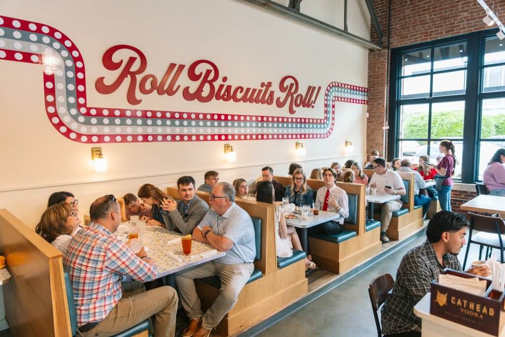 A bustling diner with patrons enjoying meals in booths, under a large 'Roll Biscuits Roll!' sign in Tuscaloosa, Alabama.