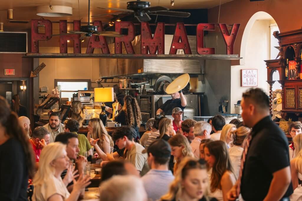 A bustling restaurant filled with people enjoying food and drinks under a large 'PHARMACY' sign in Tuscaloosa, Alabama, located at Depalma's Italian Cafe