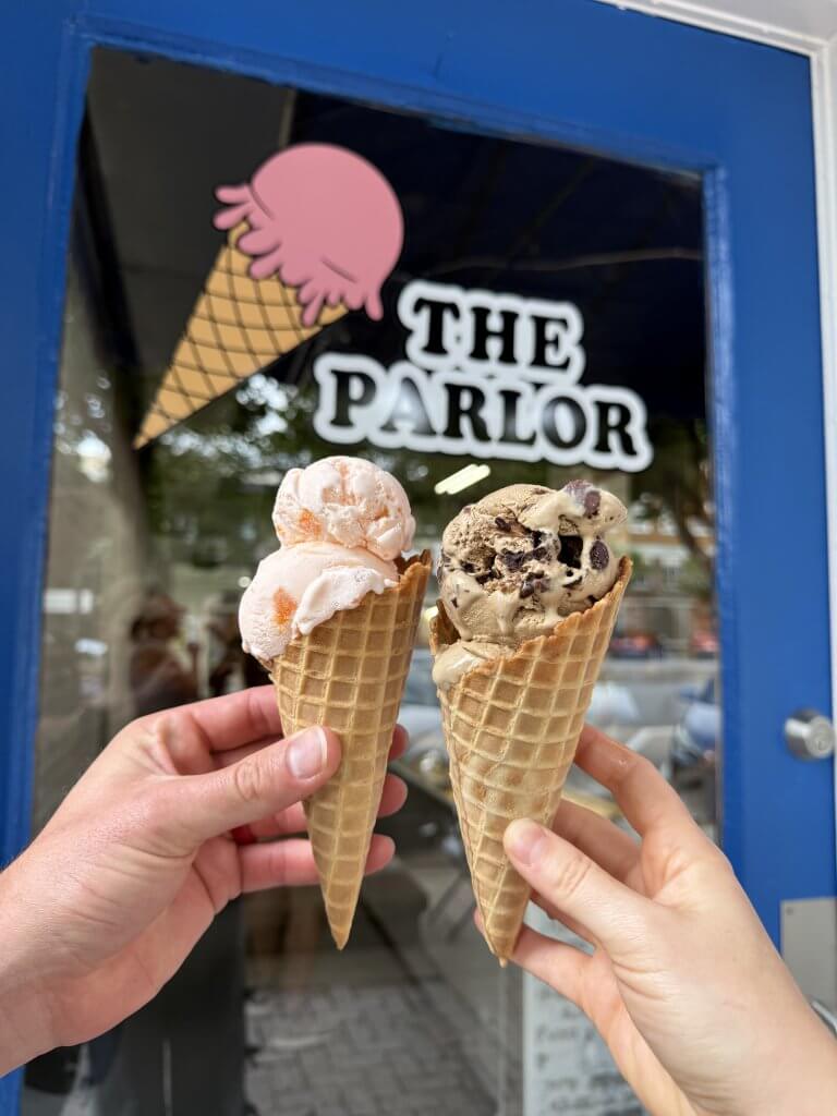 Two hands holding waffle cones with scoops of ice cream in front of an ice-cream parlor in Tuscaloosa, Alabama.