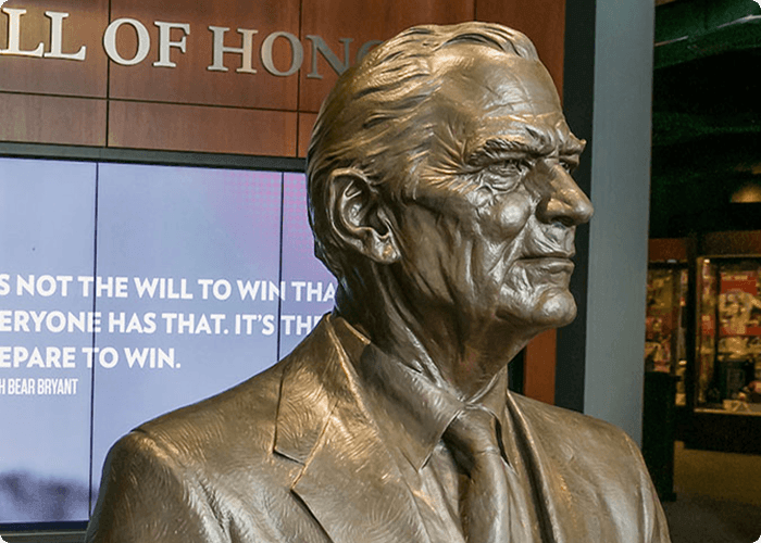 A bronze statue of an elderly man in a suit, standing in front of a screen displaying a motivational quote in Tuscaloosa, Alabama.