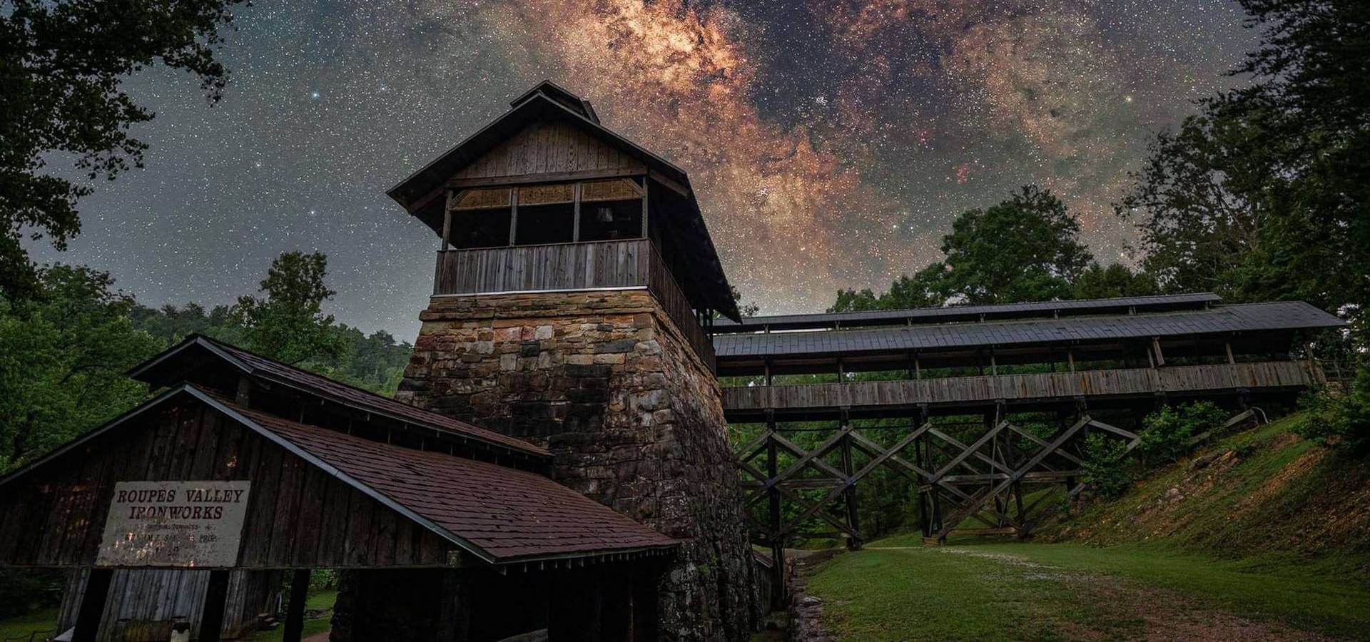 A rustic wooden tower and bridge stand under a breathtaking starry sky with the Milky Way glowing above a lush forest at Tannehill Ironworks Historical State Park, located outside of Tuscaloosa, Alabama