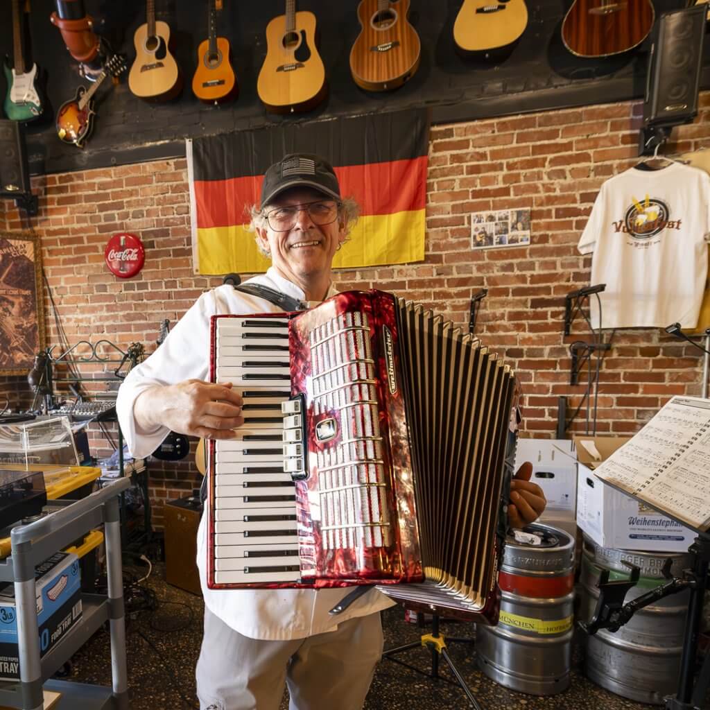 A musician stands in a music shop filled with guitars, holding a vibrant red accordion and smiling warmly in Tuscaloosa, Alabama.