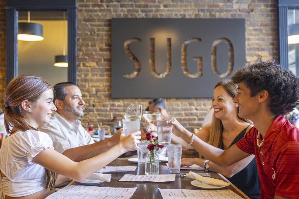 A family of four and two friends raise their glasses in a toast at a restaurant with a brick wall and the word 'SUGO' displayed prominently in Tuscaloosa, Alabama.