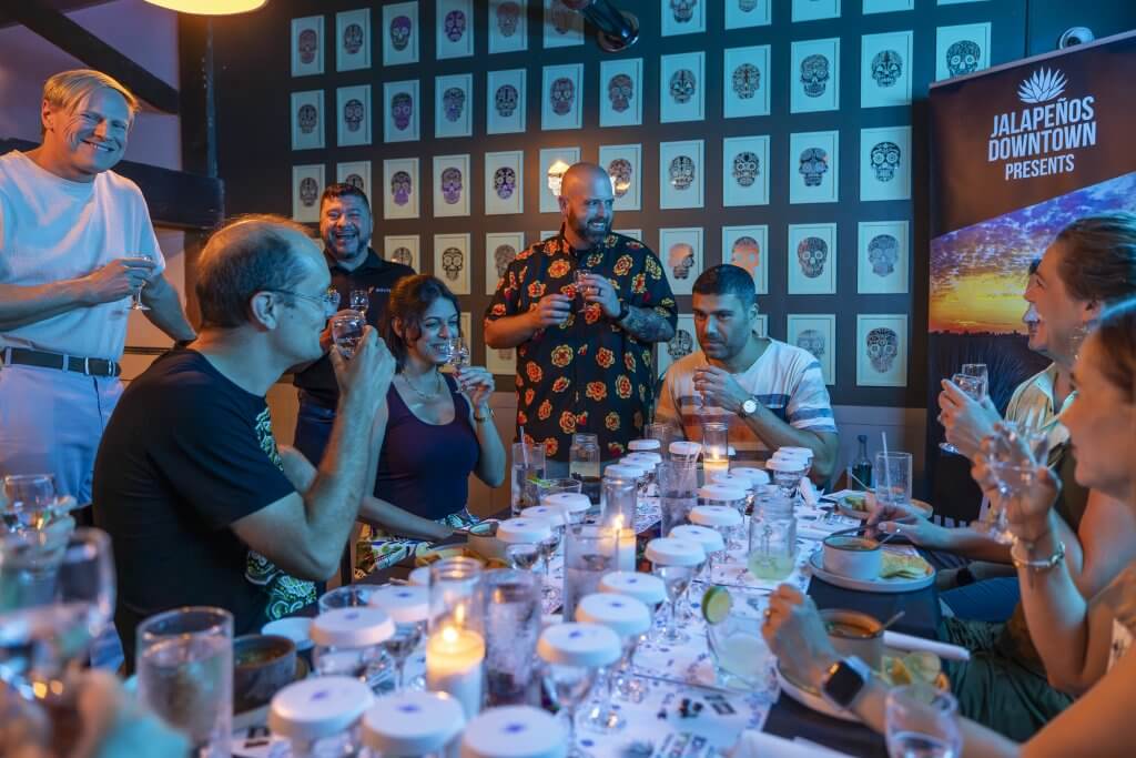 A lively group of people enjoys a festive dinner party with tequila tasting, surrounded by colorful sugar skull artwork in Tuscaloosa, Alabama.