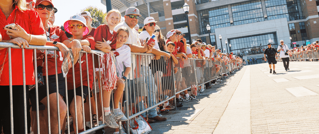 A crowd of enthusiastic fans, many wearing red, line a barricaded street, eagerly awaiting a parade or event, with a modern building in the background in Tuscaloosa, Alabama.