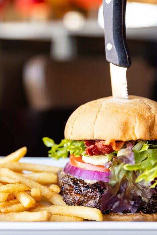 A mouthwatering burger with lettuce, tomato, onion, and bacon, topped with a knife, served with crispy fries on the side in Tuscaloosa, Alabama.