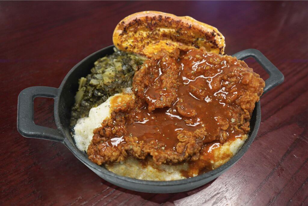 A hearty serving of chicken smothered in gravy, accompanied by mashed potatoes, collard greens, and a piece of cornbread in a cast-iron skillet in Tuscaloosa, Alabama.