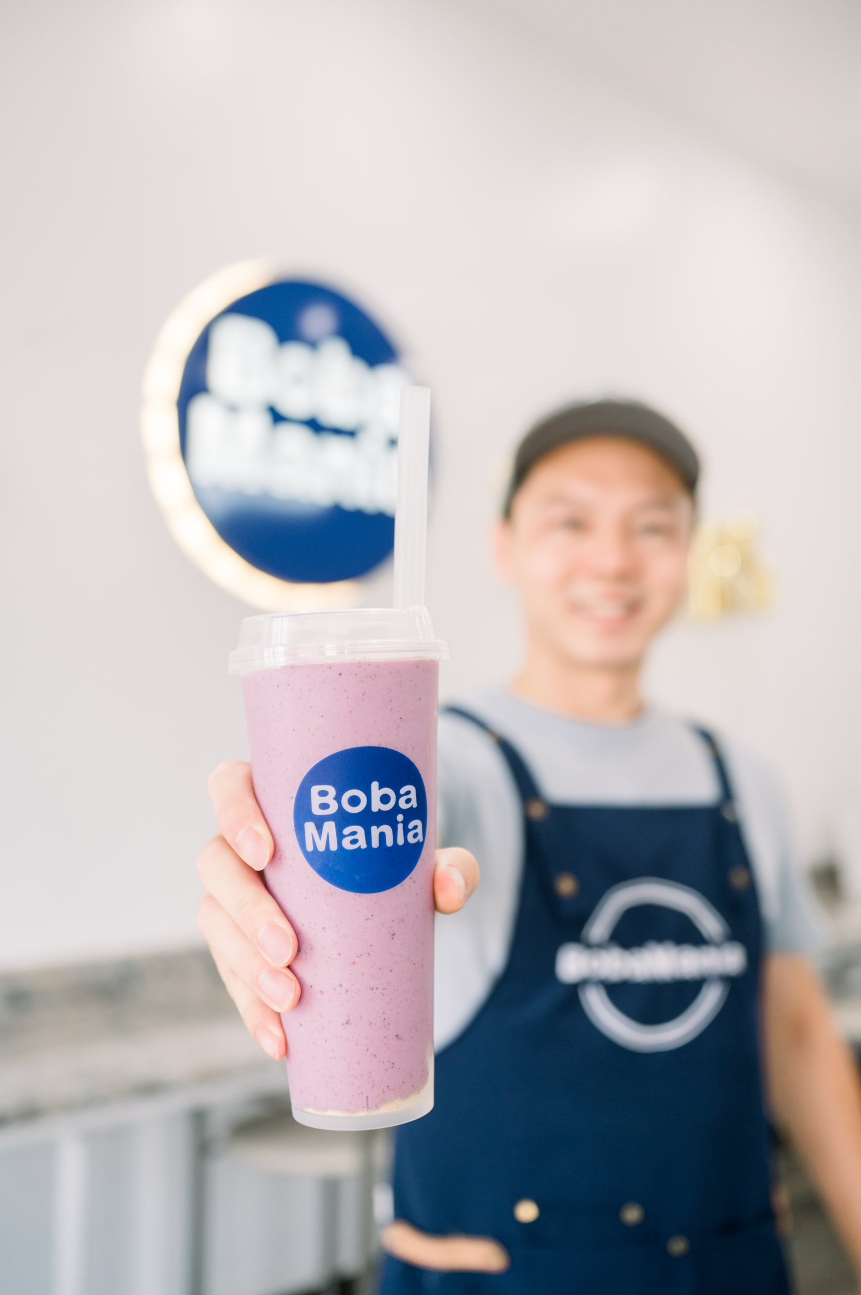A smiling barista proudly holds up a vibrant purple bubble tea from Boba Mania, with the store's logo prominently displayed in the background in Tuscaloosa, Alabama.