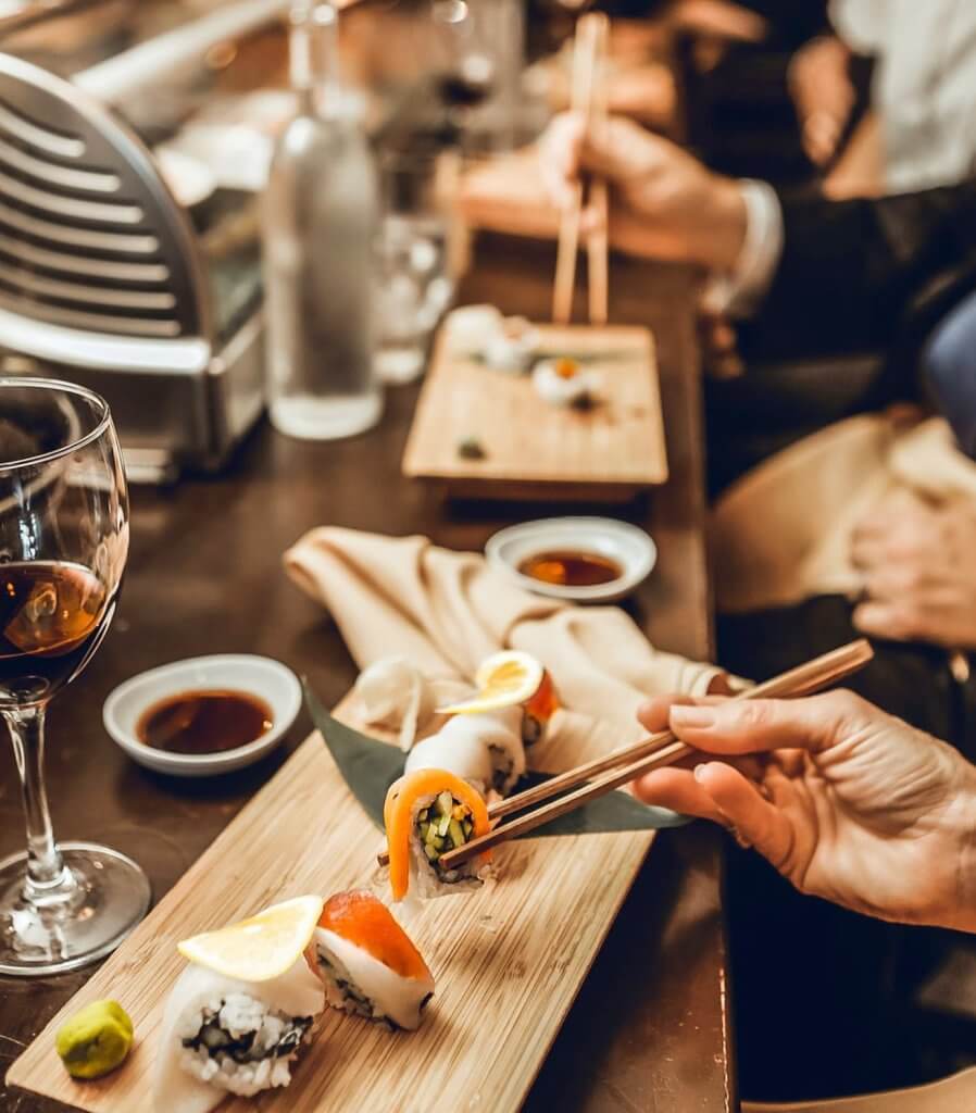 A person skillfully uses chopsticks to pick up a piece of sushi from a beautifully arranged wooden platter, surrounded by a glass of red wine, soy sauce, and wasabi in Tuscaloosa, Alabama.