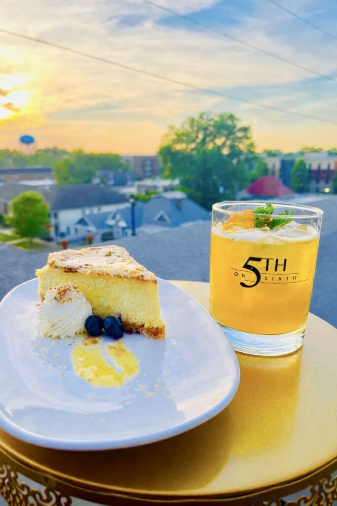 A slice of creamy cheesecake garnished with whipped cream and blueberries, served on a white plate next to a glass of refreshing yellow cocktail with a mint sprig, both placed on a golden table with a stunning sunset view of a cityscape in the background in Tuscaloosa, Alabama.