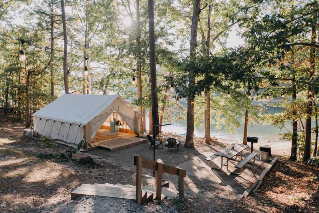 A serene lakeside campsite with a spacious white tent, wooden picnic tables, and a fire pit nestled among tall trees in Tuscaloosa, Alabama.